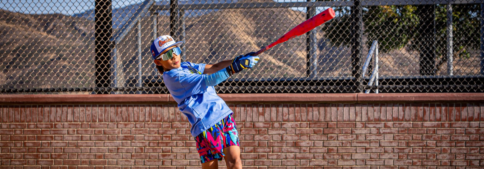 Person playing baseball in a batting stance with a red bat, wearing a blue shirt and colorful shorts.
