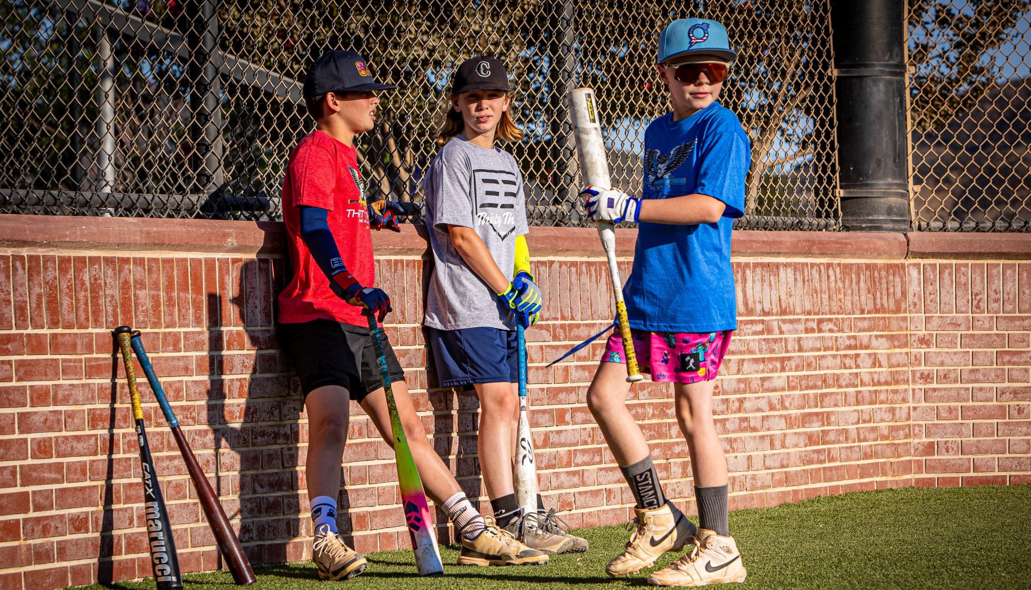 Three children with baseball bats leaning against a brick wall.