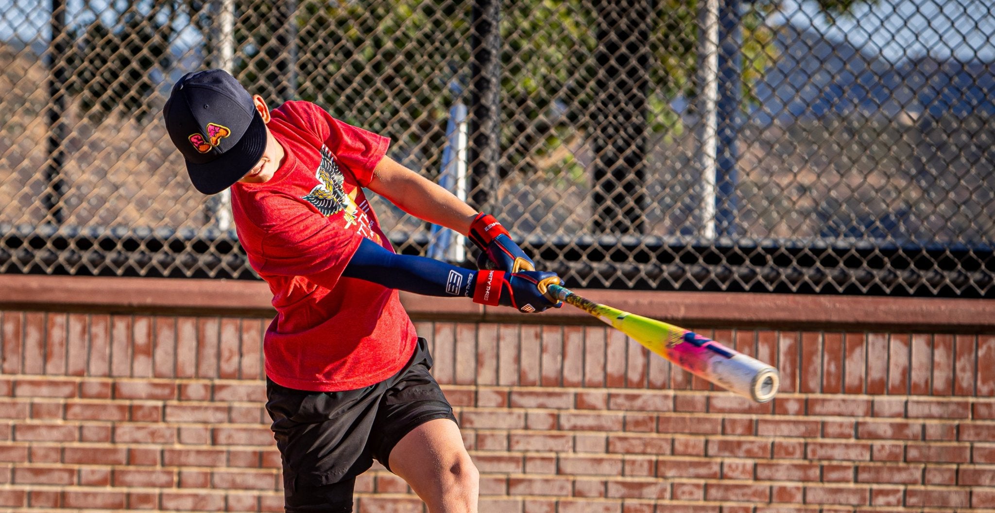 Person in red shirt and black cap swinging a colorful baseball bat against a chain-link fence background.