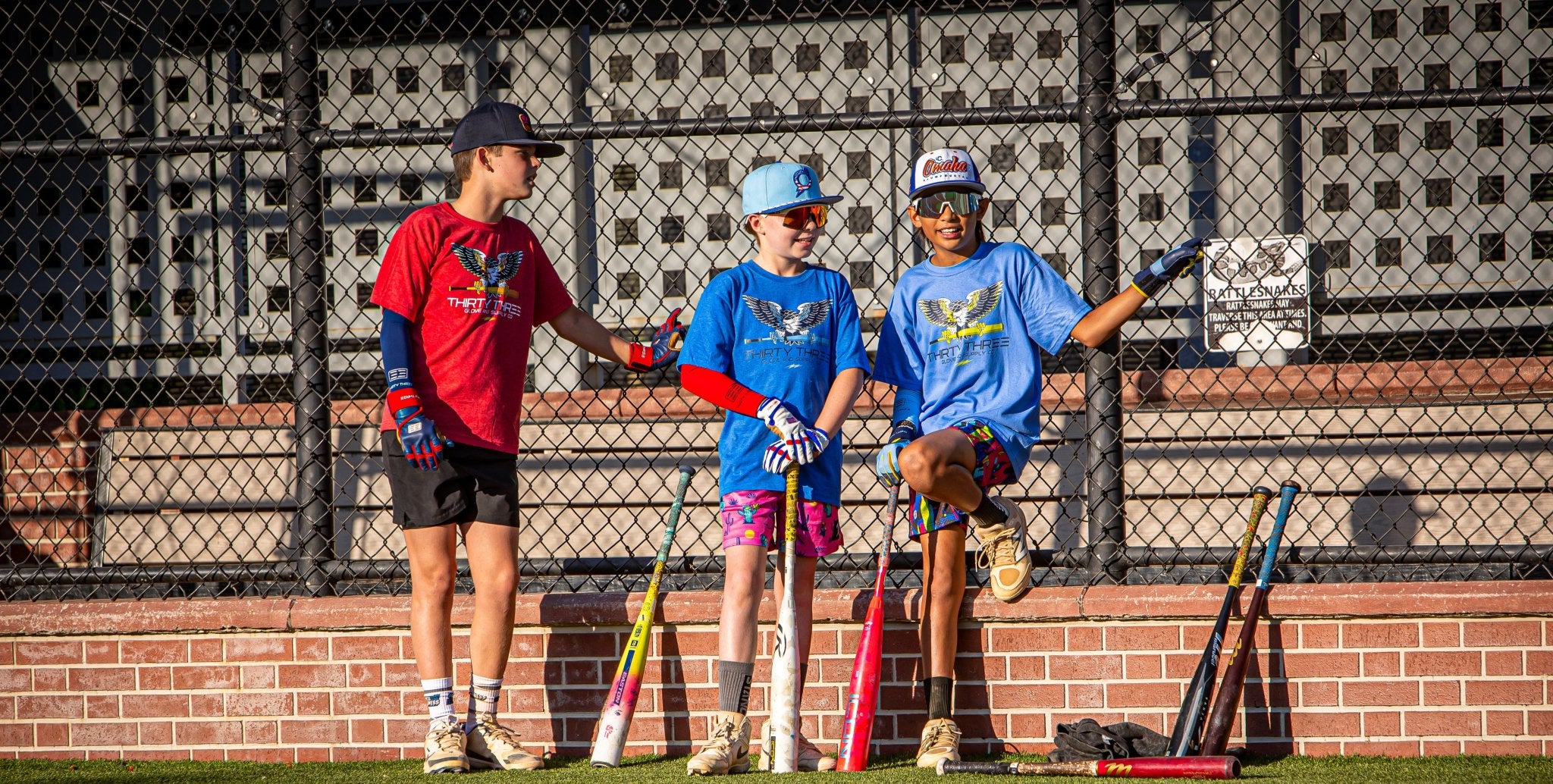 Three children in sports attire standing on a baseball field with bats.