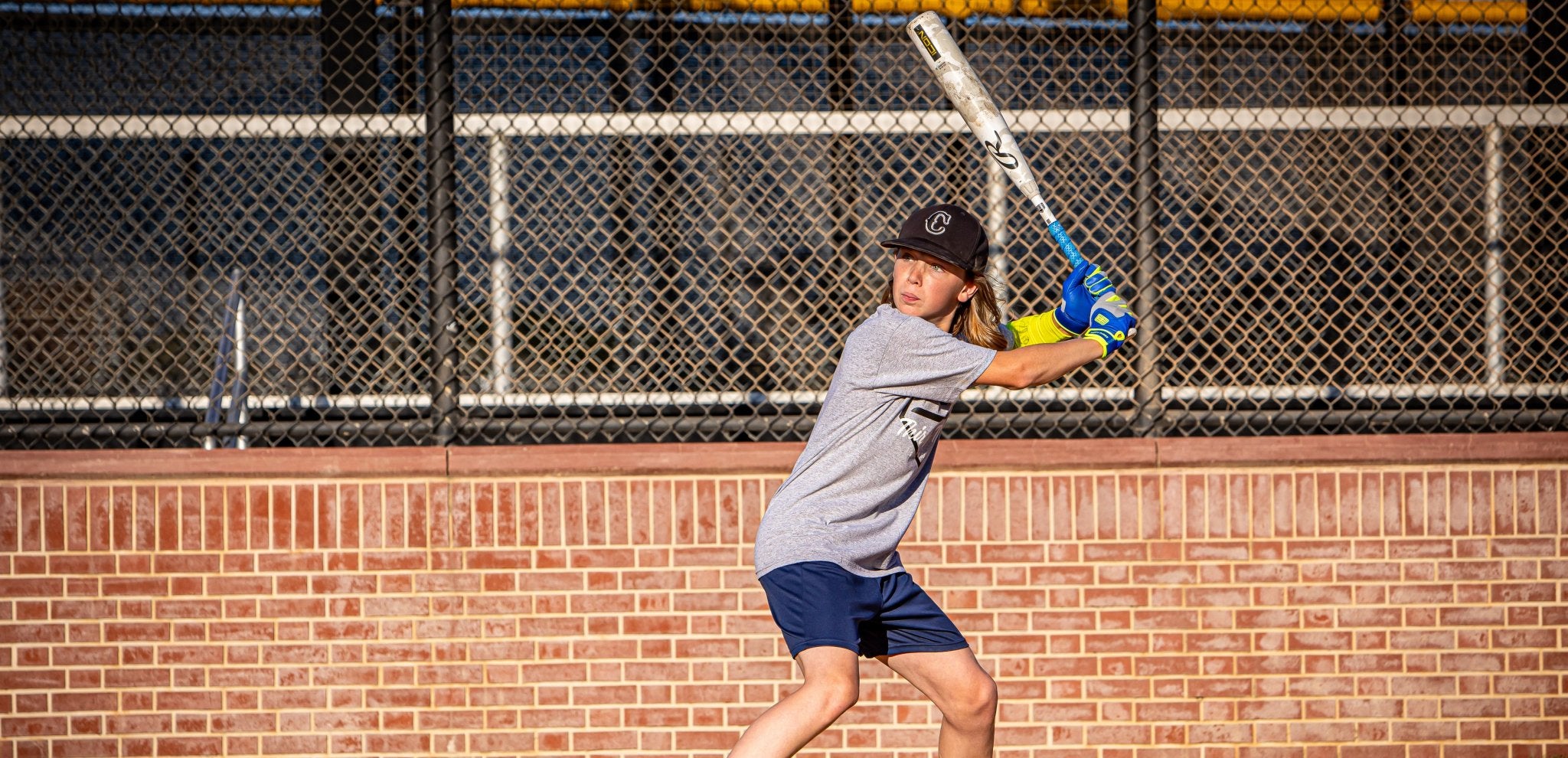 Person playing baseball or softball in front of a chain-link fence and brick wall.