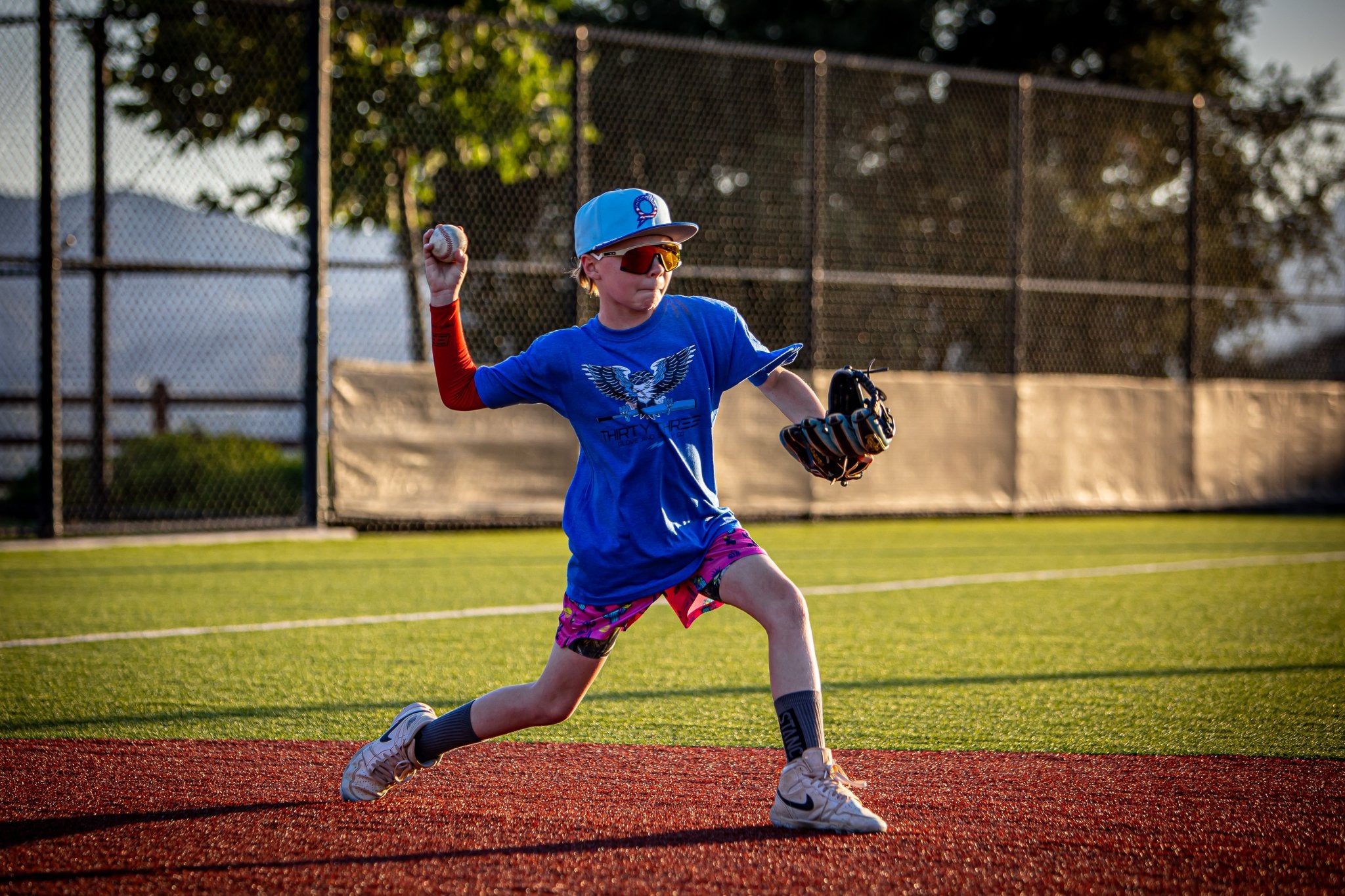 Child in a blue baseball uniform pitching a ball on a field with trees and a fence in the background.